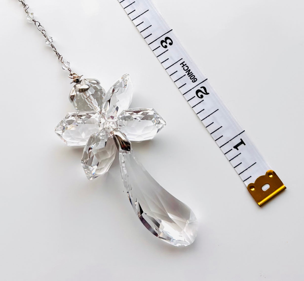 A clear faceted crystal fairy angel showing a teardrop-shaped drop, shown on a white background next to a measuring tape for scale.