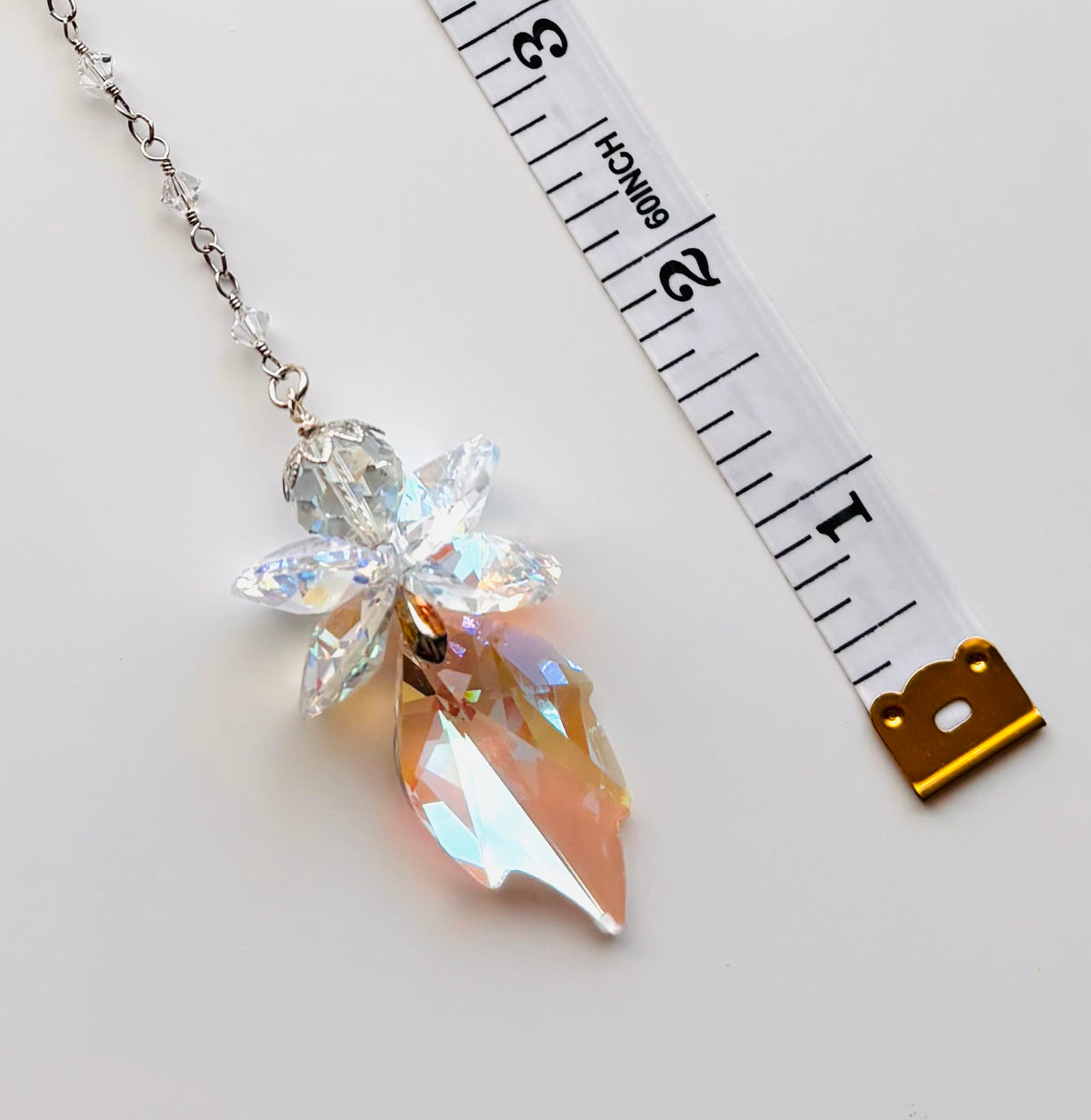 A crystal fairy pendant necklace with a silver chain, shown next to a white measuring tape to indicate scale on a light gray background.