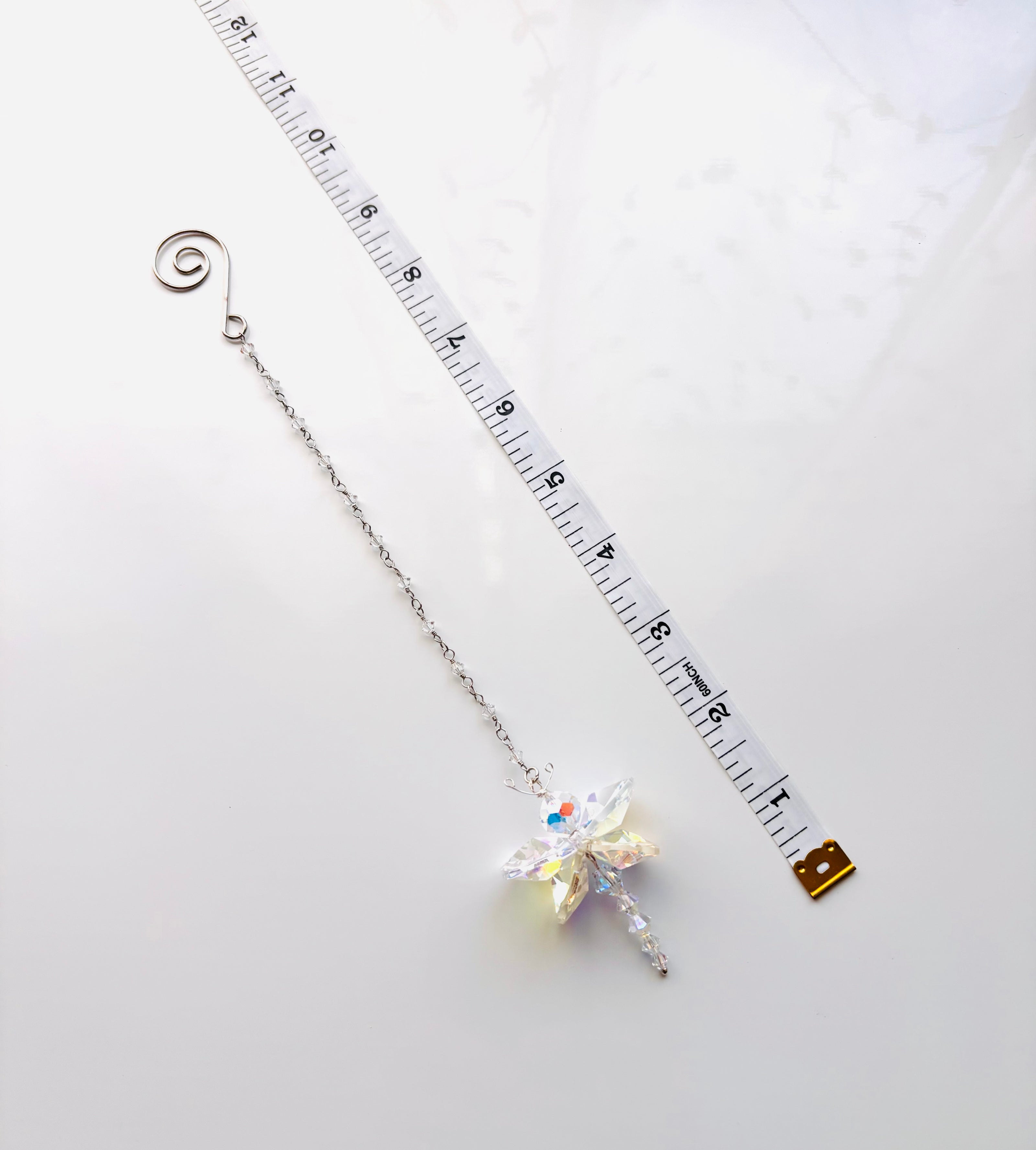 A silver chain accessory with a spiral clasp and a dangling iridescent butterfly charm, shown on a white background with a measuring tape and small gold tag nearby for scale.