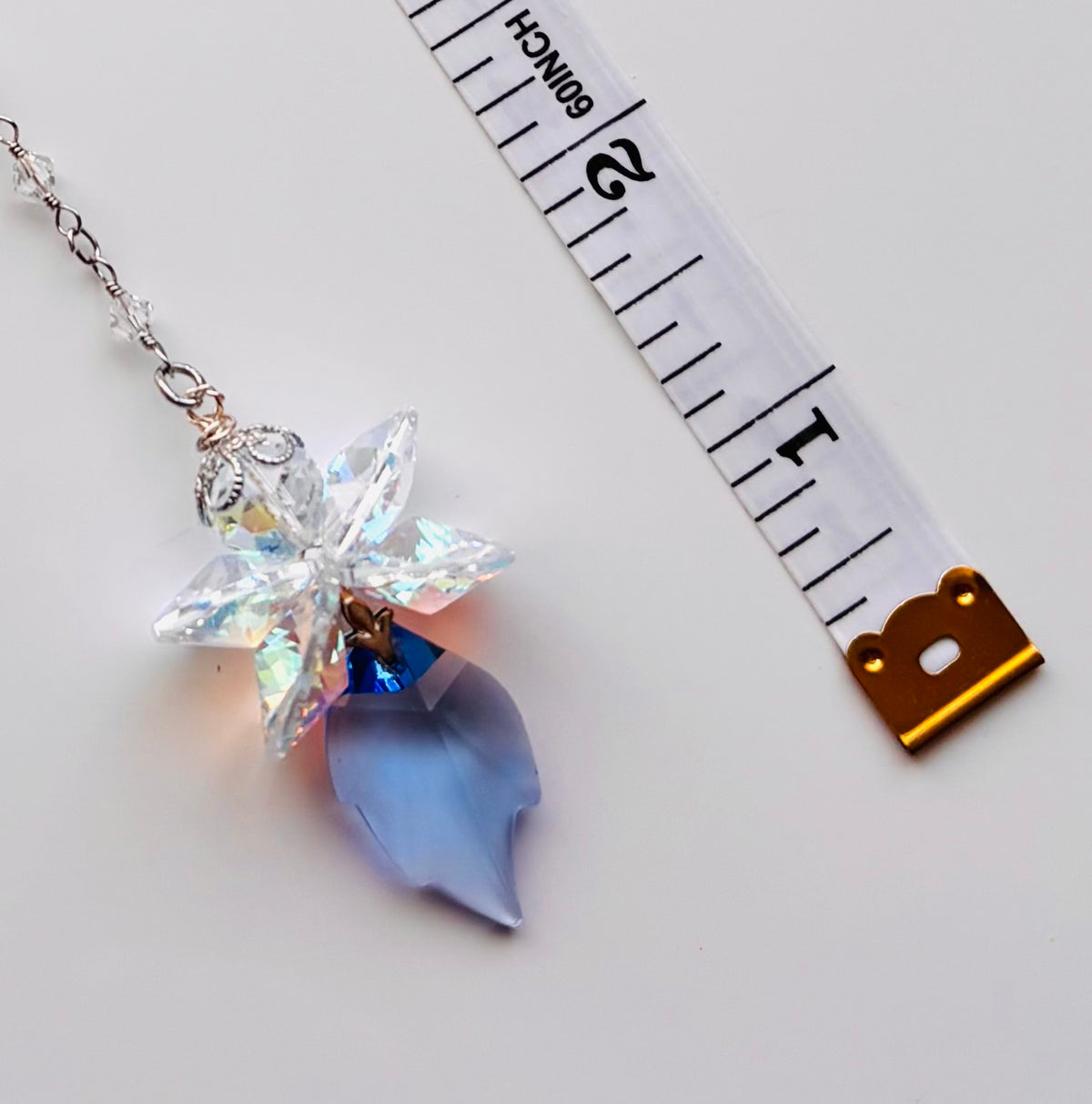 A crystal fairy pendant necklace with a faceted clear top, an iridescent blue flower, and a light blue teardrop charm on a silver chain, shown next to a white measuring tape for scale on a white background.