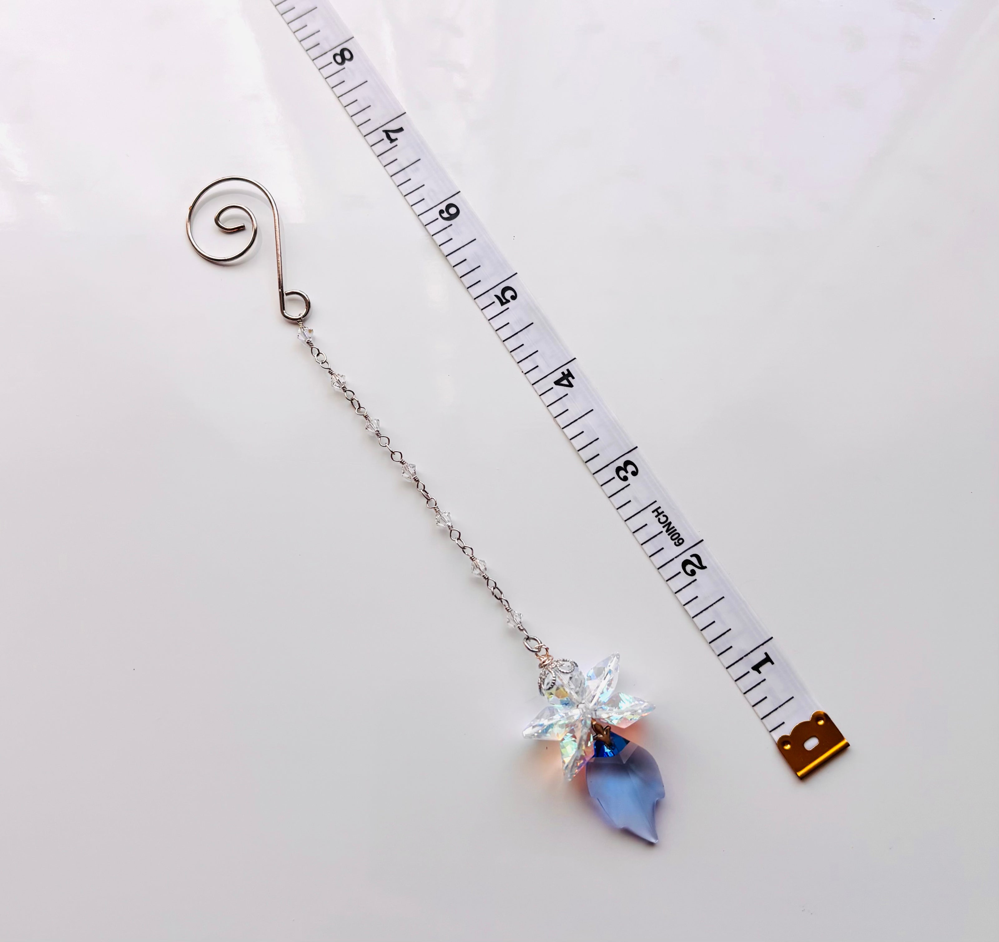 A suncatcher with a spiral end, a delicate chain, and a dangling crystal flower and blue teardrop charm, shown next to a measuring tape for scale on a white background.