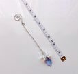 A suncatcher with a spiral end, a delicate chain, and a dangling crystal flower and blue teardrop charm, shown next to a measuring tape for scale on a white background.