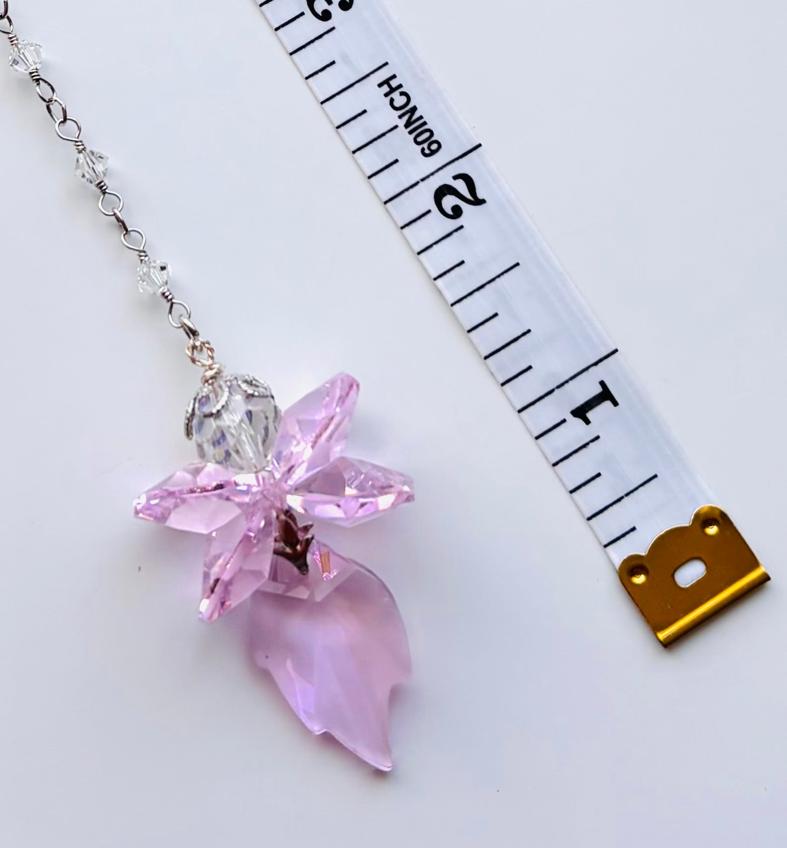 A pink crystal fairy pendant necklace on a silver chain, shown next to a white measuring tape to indicate scale, on a clean white background.