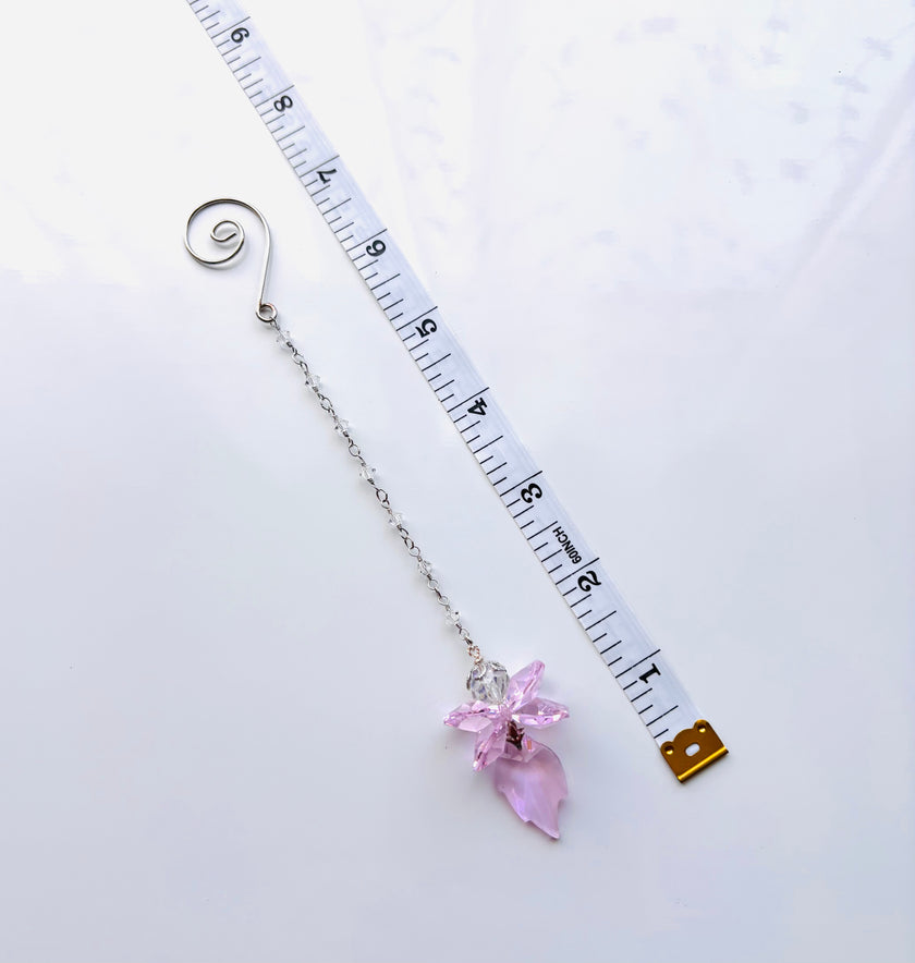 A silver chain with a pink crystal flower fairy pendant and a spiral end, shown next to a white measuring tape on a white background.