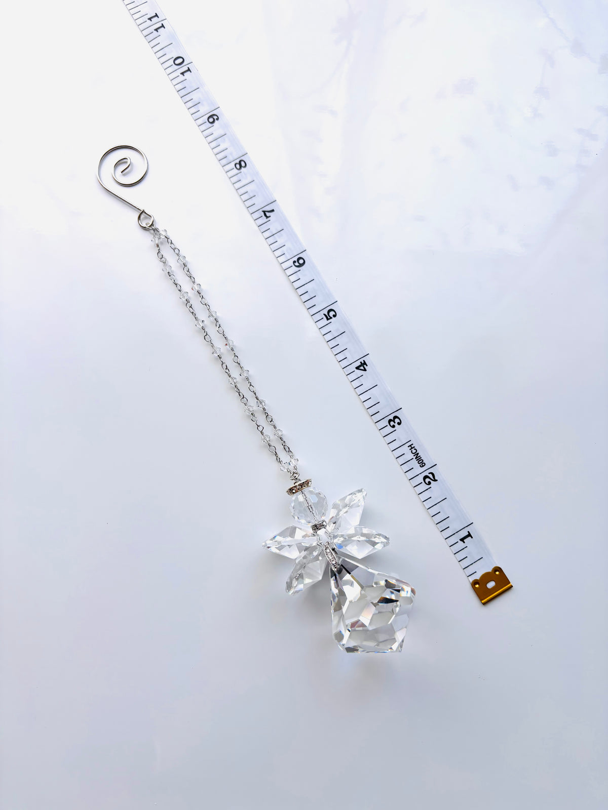A silver-toned chain accessory with a clear faceted crystal angel pendant and a spiral ring at the top, shown on a white background next to a measuring tape for scale.