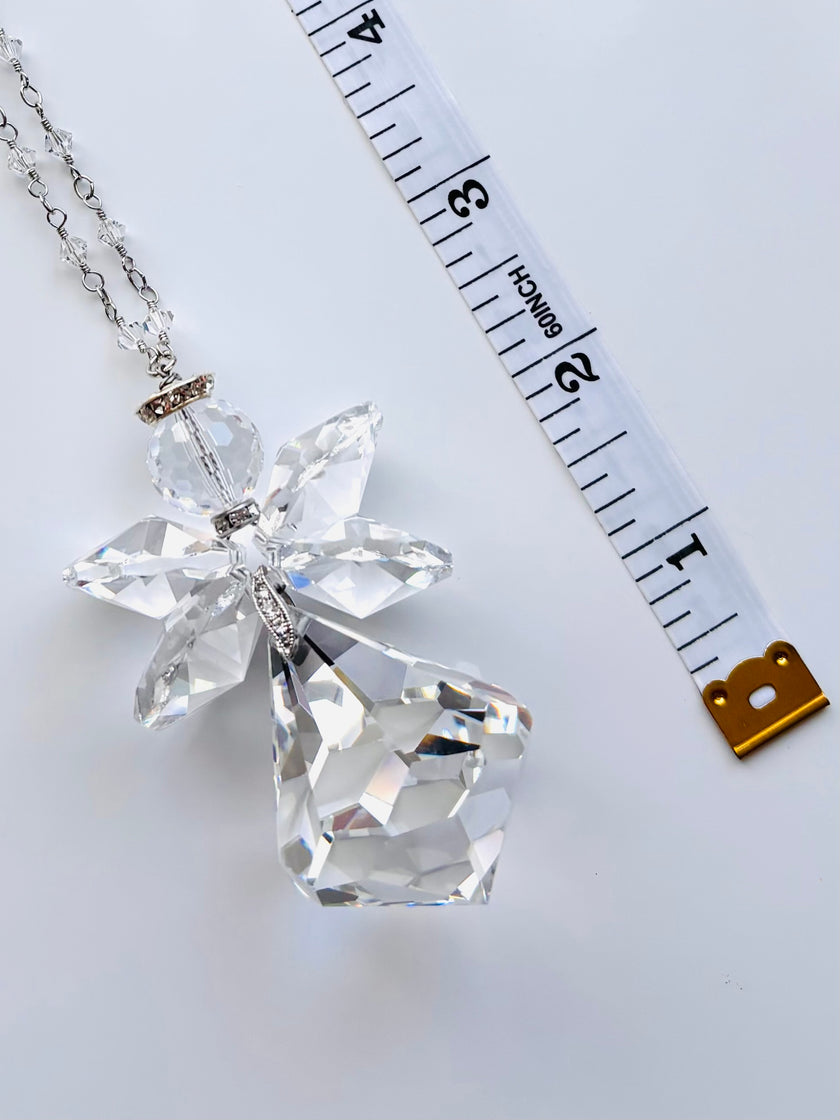 A clear faceted crystal angel pendant necklace with a silver-toned chain, displayed next to a white measuring tape to show scale on a light gray background.