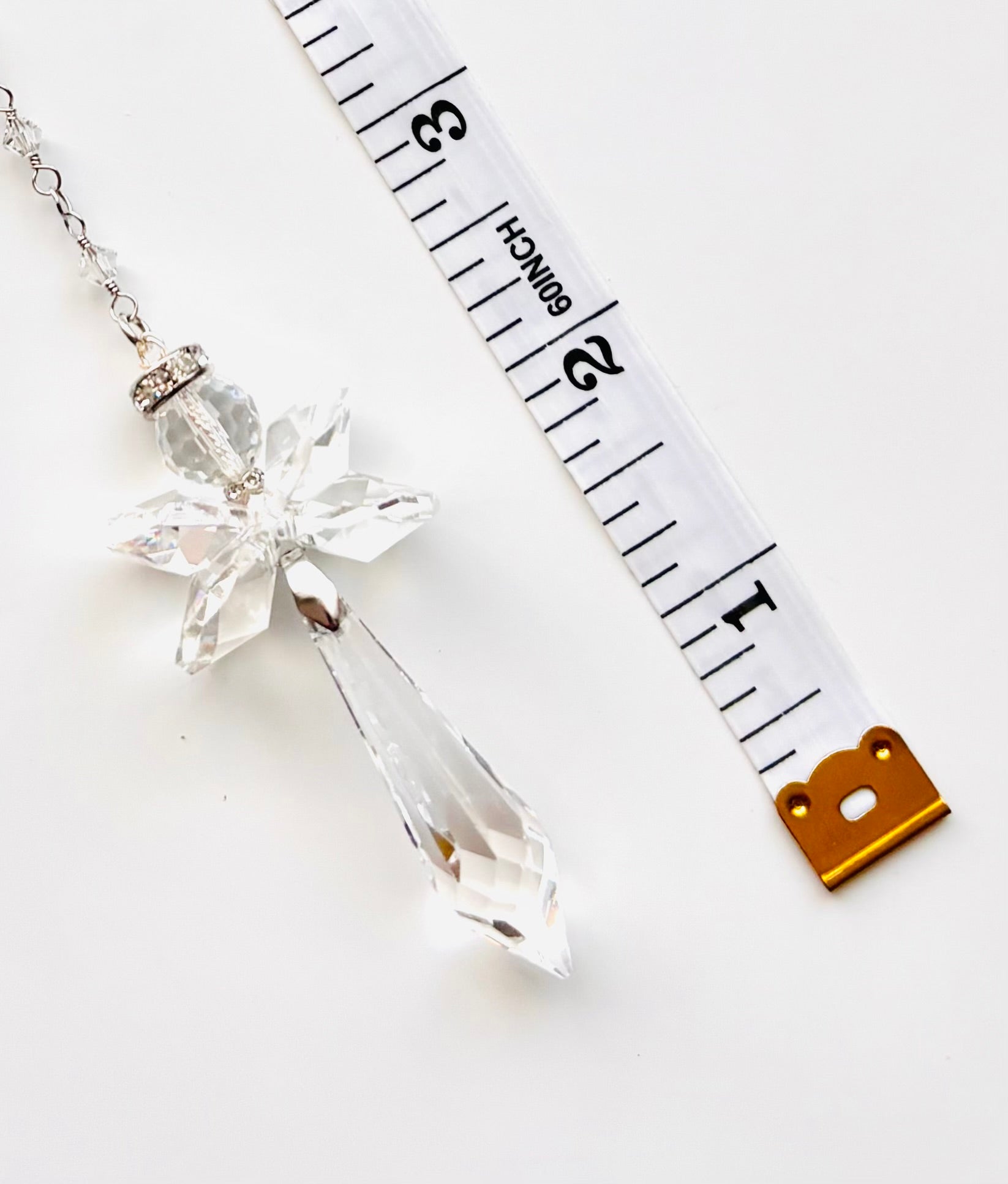 A clear faceted crystal angel pendant necklace with a silver-toned chain and cap, shown next to a white measuring tape to indicate scale on a white background.
