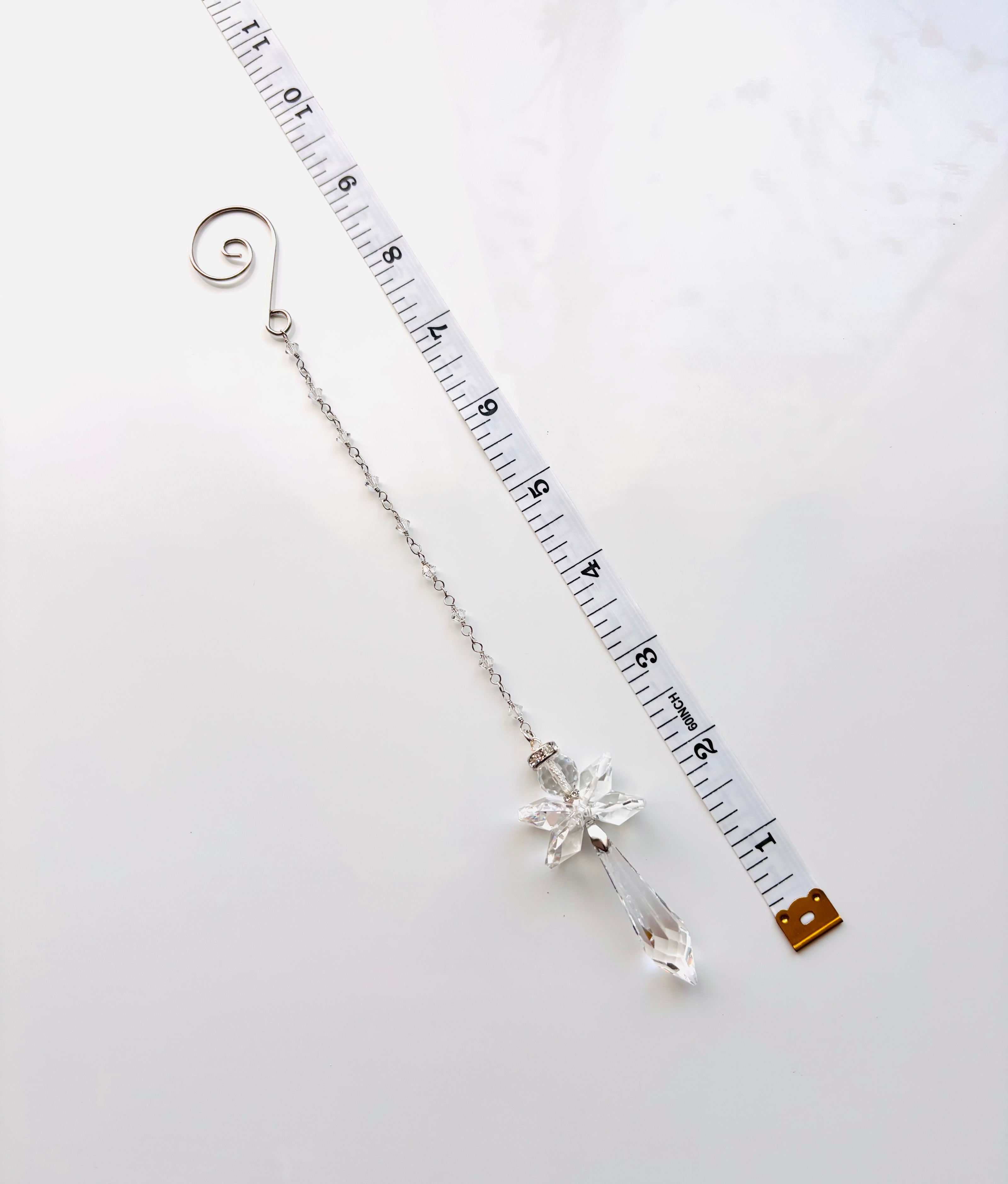 A silver-toned  beaded chain with a clear crystal angel drop pendant, shown next to a white measuring tape to indicate its length on a light gray background.