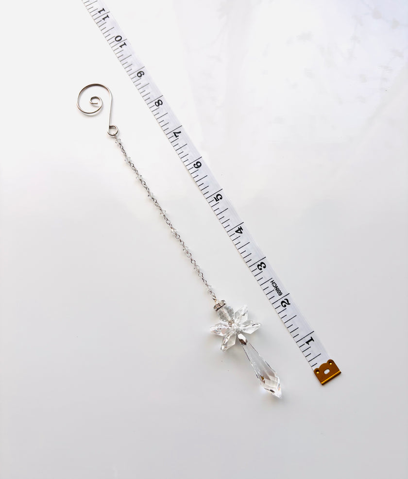 A silver-toned  beaded chain with a clear crystal angel drop pendant, shown next to a white measuring tape to indicate its length on a light gray background.