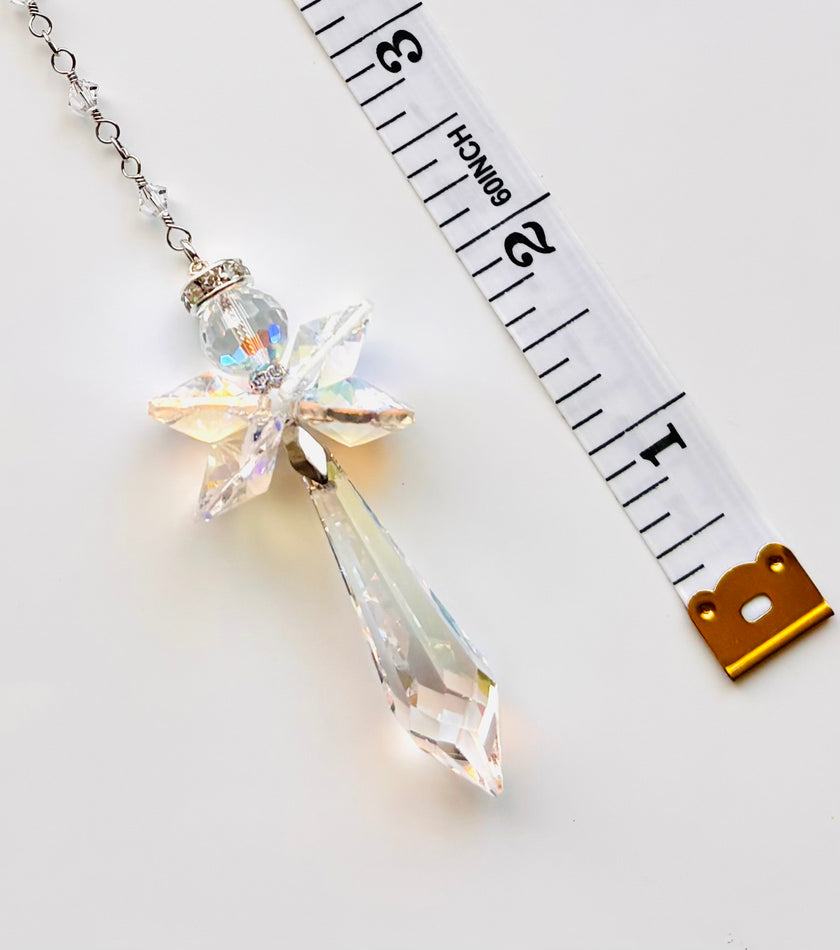 A clear iridescent crystal angel pendant necklace on a silver chain, shown next to a white measuring tape to indicate scale on a white background.