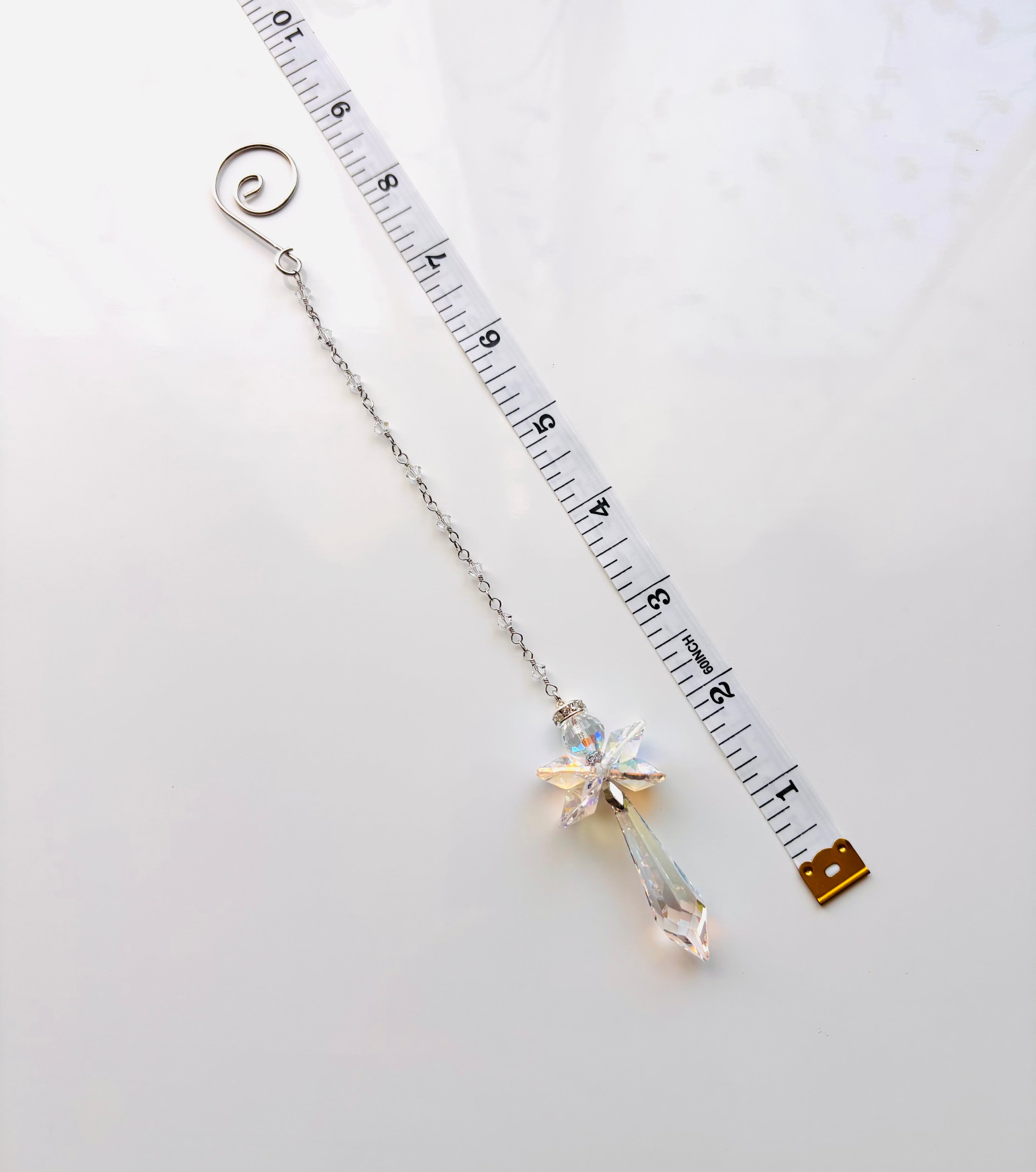 A silver-toned crystal angel with a spiral end and a dangling faceted crystal charm, shown next to a white measuring tape on a light background.