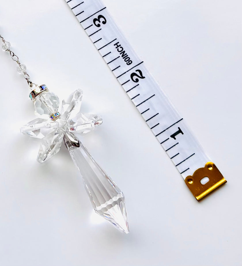 A clear crystal angel pendant necklace with a faceted crystal body and a silver-toned chain, shown next to a white measuring tape to indicate scale on a white background.