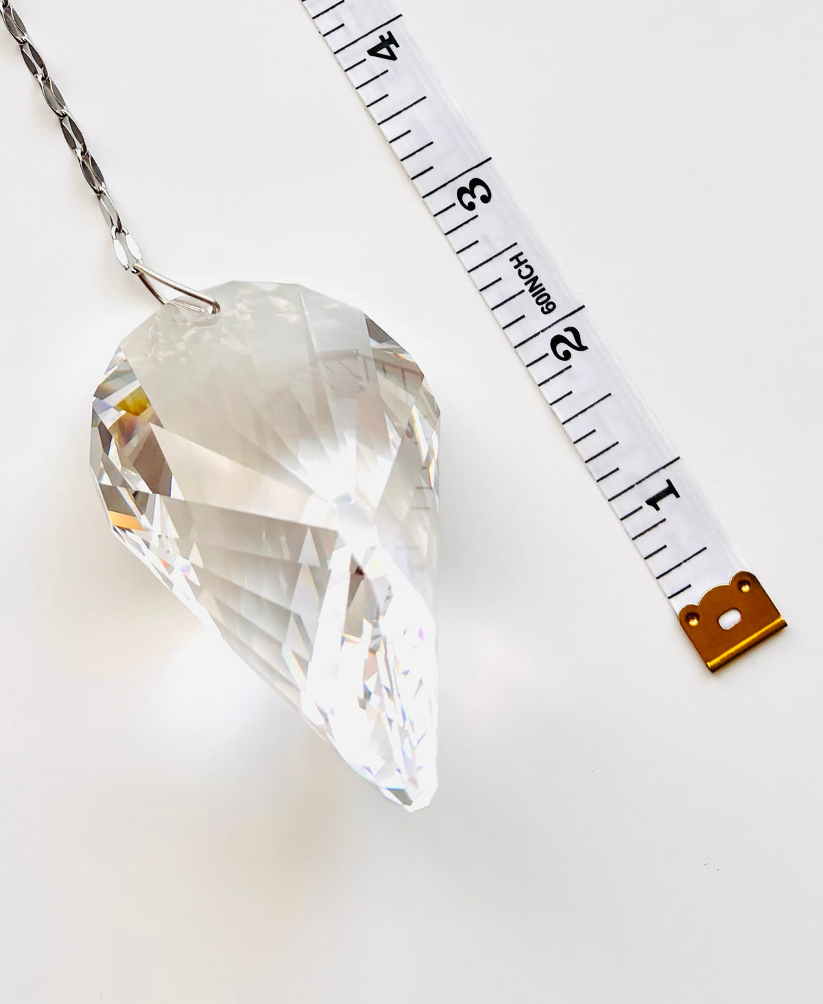 A faceted clear crystal on a silver chain, shown next to a white measuring tape to indicate size, on a clean white background.