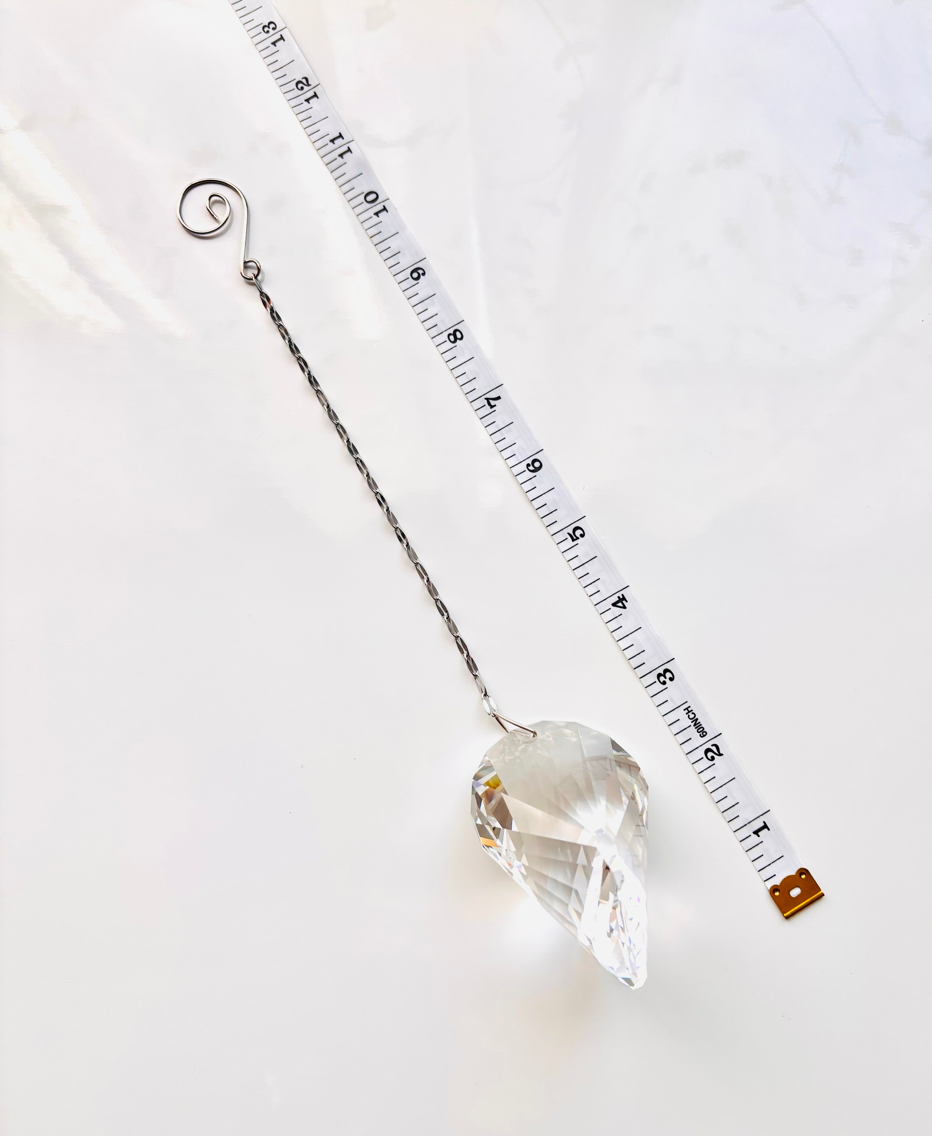 A clear faceted crystal ornament with a silver wire hanger and spiral top, shown on a white marble surface next to a measuring tape for scale.