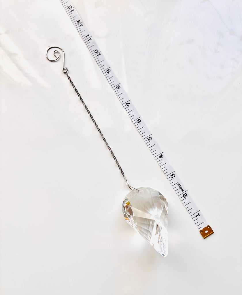 A clear faceted crystal ornament with a silver wire hanger and spiral top, shown on a white marble surface next to a measuring tape for scale.