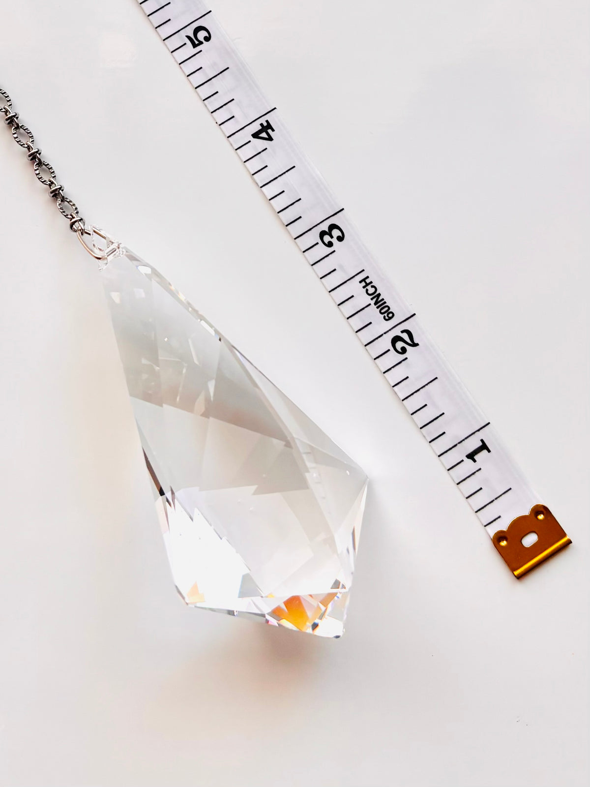 A faceted clear crystal pendant or chandelier drop shown on a white background next to a measuring tape to indicate size.