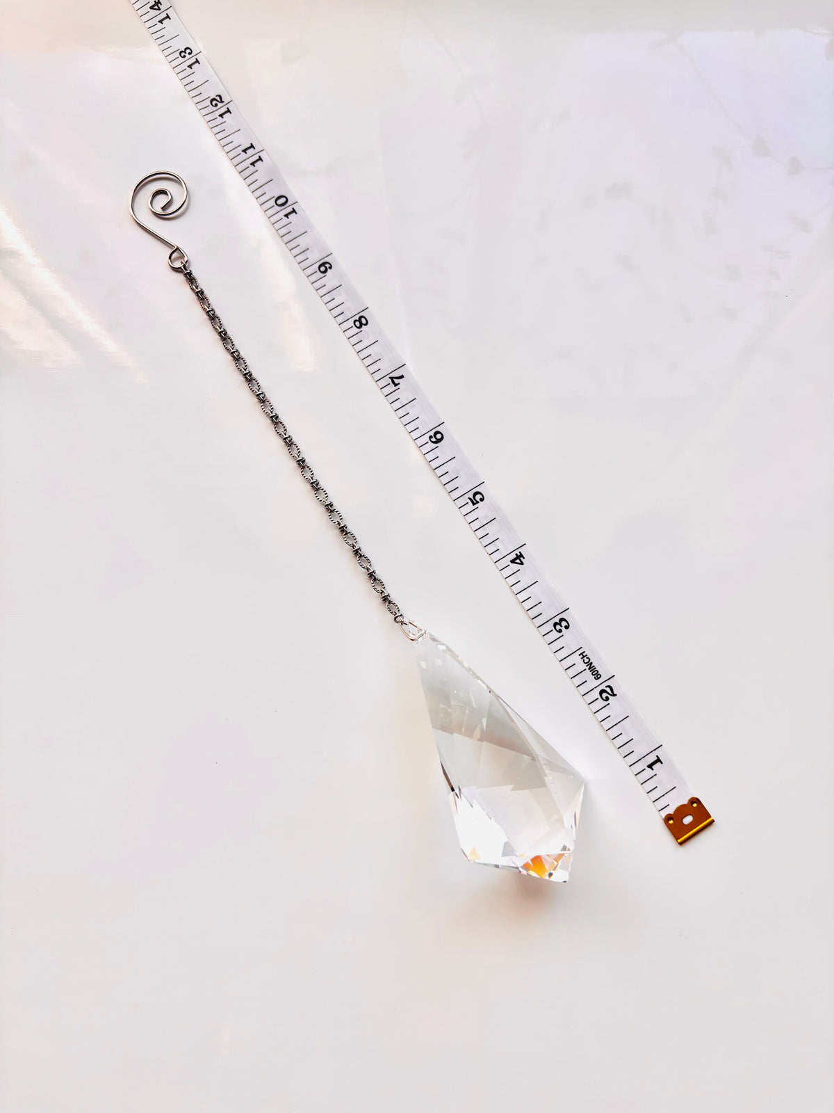 A clear faceted crystal suncatcher with a silver chain and spiral top, shown next to a measuring tape to indicate its size on a white background.