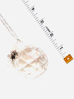 A faceted clear crystal pendant on a silver-toned chain, shown on a white background with a measuring tape and small gold tag prop.