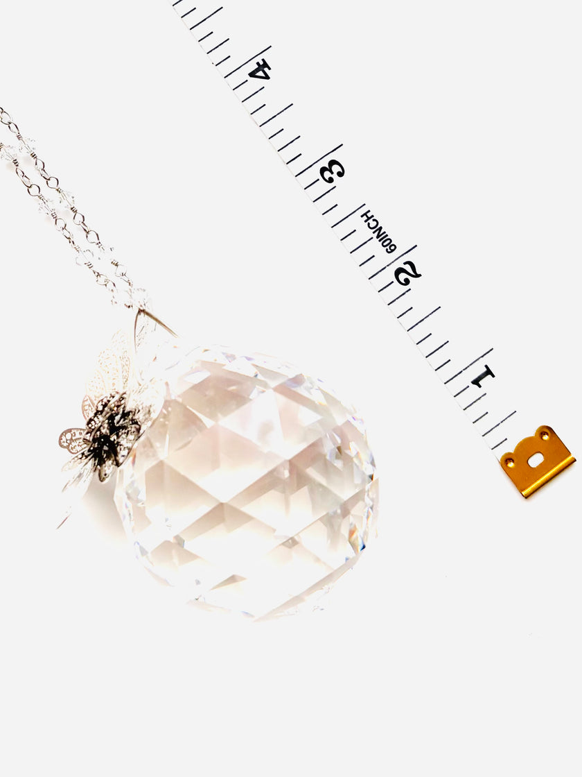 A faceted clear crystal pendant on a silver-toned chain, shown on a white background with a measuring tape and small gold tag prop.