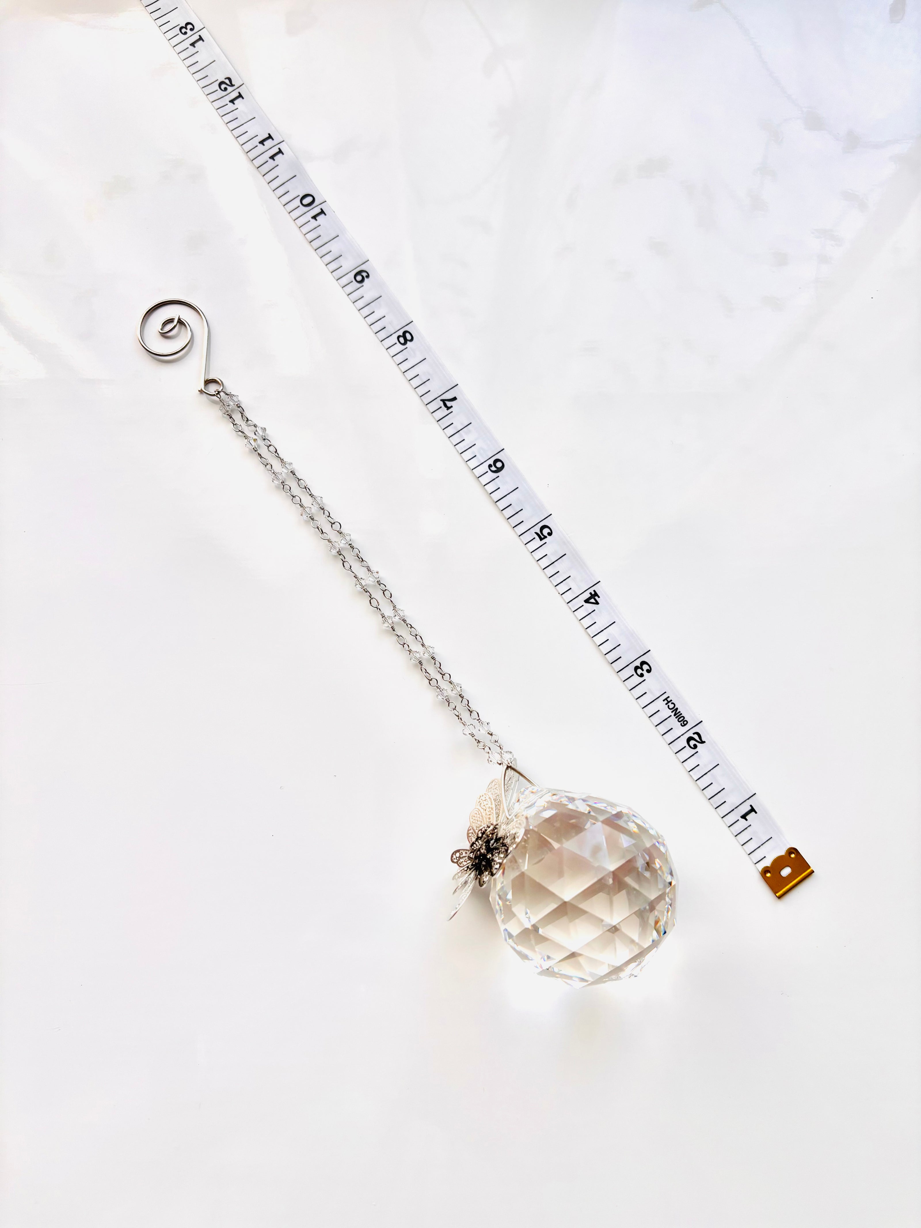A crystal ball pendant necklace with a silver chain and spiral clasp, displayed diagonally next to a measuring tape on a white marble surface.