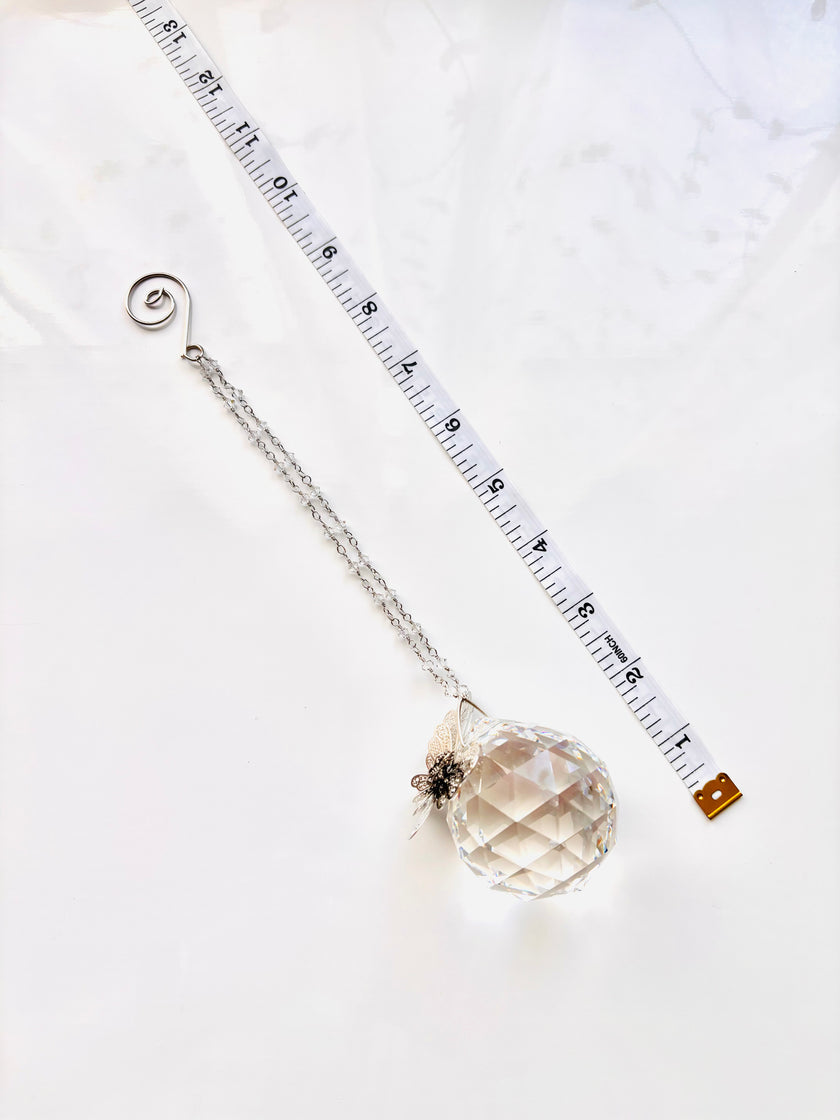 A crystal ball pendant necklace with a silver chain and spiral clasp, displayed diagonally next to a measuring tape on a white marble surface.