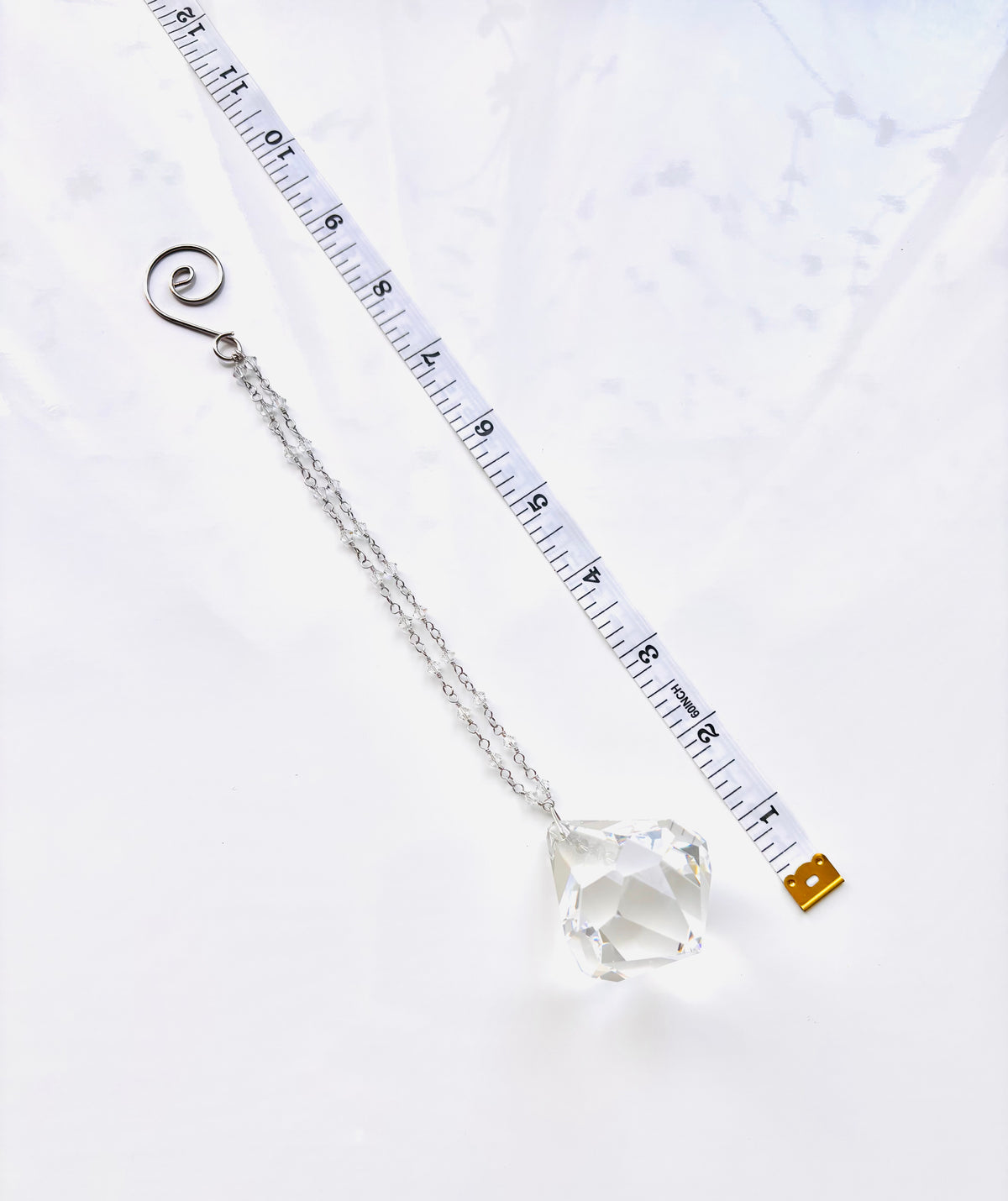 A silver chain with a faceted clear crystal pendant and a spiral clasp, laid diagonally next to a white measuring tape on a white background.