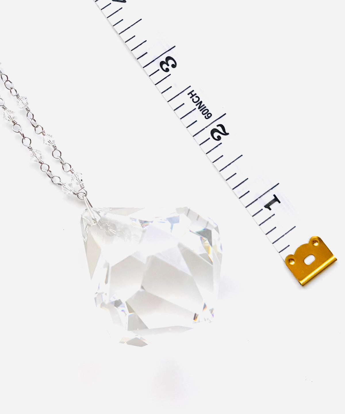 A faceted clear crystal pendant on a silver chain, shown next to a measuring tape and a small gold scale icon on a white background.