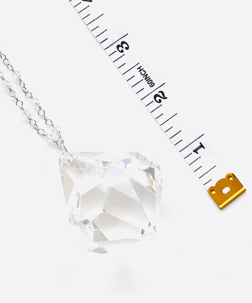 A faceted clear crystal pendant on a silver chain, shown next to a measuring tape and a small gold scale icon on a white background.
