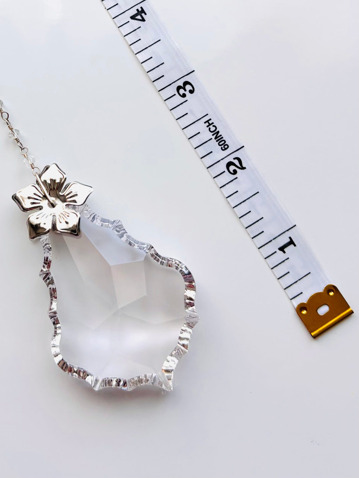 A silver-toned pendant with a faceted clear crystal drop and a silver flower clasp, shown next to a white measuring tape on a white background.