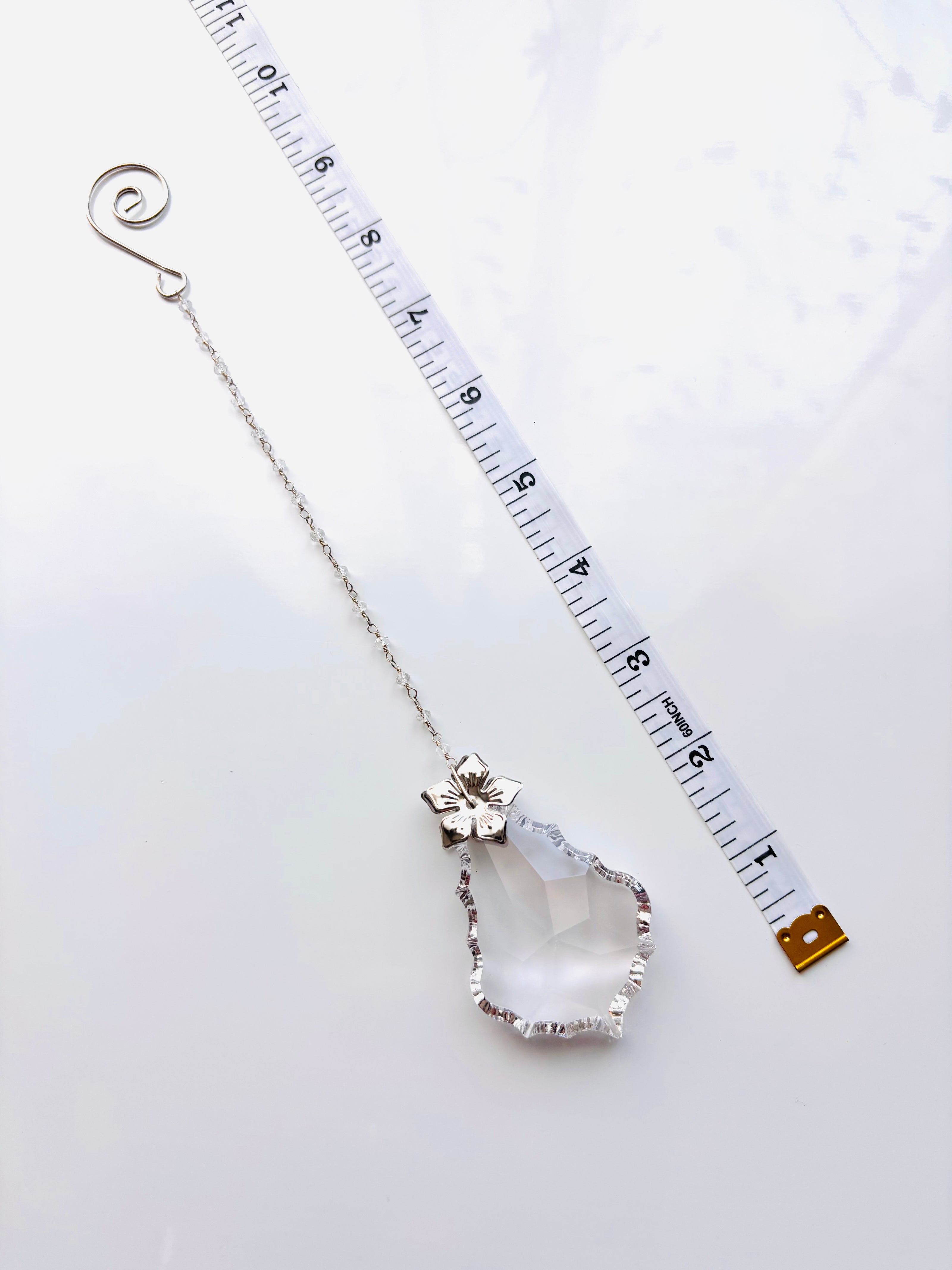 A silver-toned chain with a large baroque, faceted clear crystal pendant, displayed next to a white measuring tape to show scale on a white background.