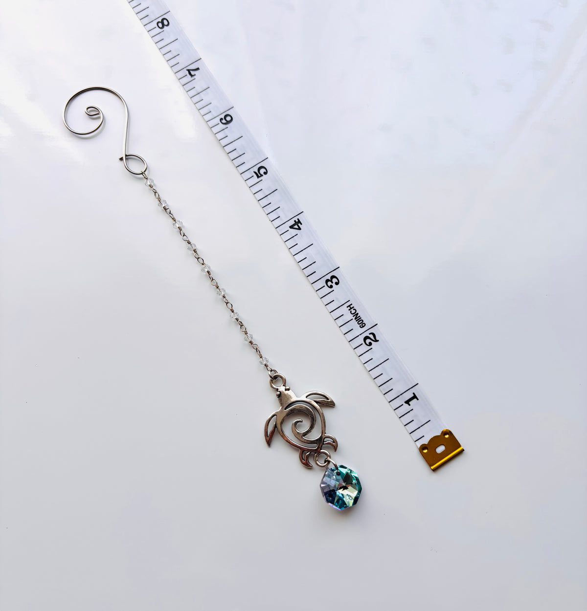 A silver-toned turtle with a decorative swirl design and a small iridescent blue-green faceted crystal pendant, shown next to a white measuring tape on a white background.