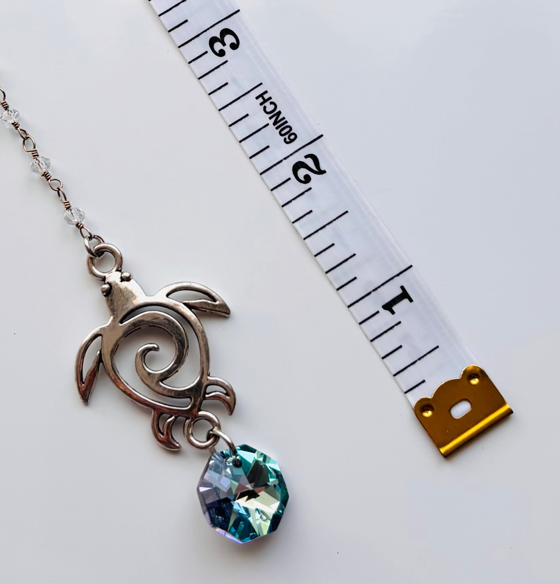 A silver-toned turtle pendant charm with a faceted blue-green crystal drop, shown next to a white measuring tape for scale on a white background.