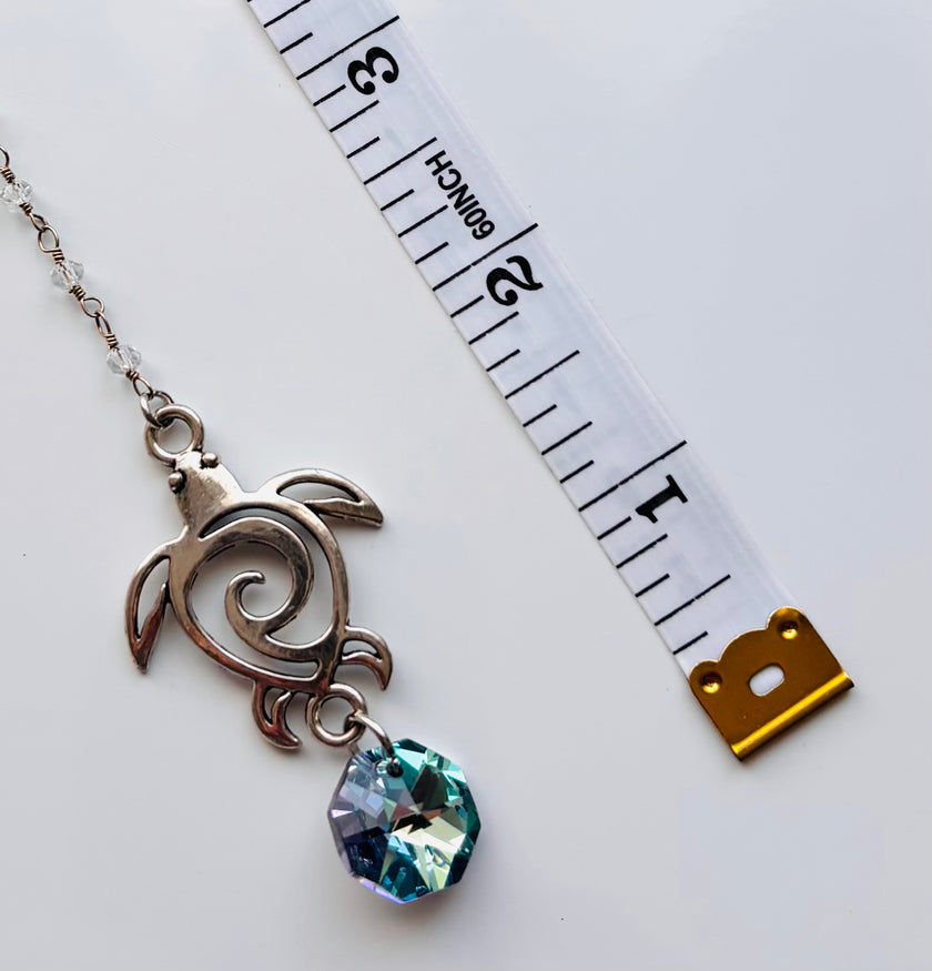 A silver-toned turtle pendant charm with a faceted blue-green crystal drop, shown next to a white measuring tape for scale on a white background.