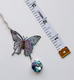 A butterfly-shaped pendant with a faceted blue crystal charm, displayed next to a white measuring tape on a white background.