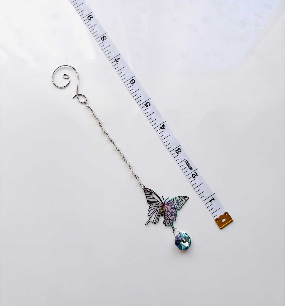 A silver-tone butterfly on beaded chain  with a dangling iridescent blue gemstone, shown next to a white measuring tape to indicate scale on a white background.