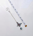 A silver-tone butterfly on beaded chain  with a dangling iridescent blue gemstone, shown next to a white measuring tape to indicate scale on a white background.