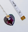 A silver heart-shaped pendant with layered angel wings and a small yellow flower charm, shown next to a measuring tape for scale on a white background.
