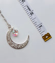 Silver crescent moon pendant with a dangling iridescent heart charm, shown next to a white measuring tape on a light gray background.