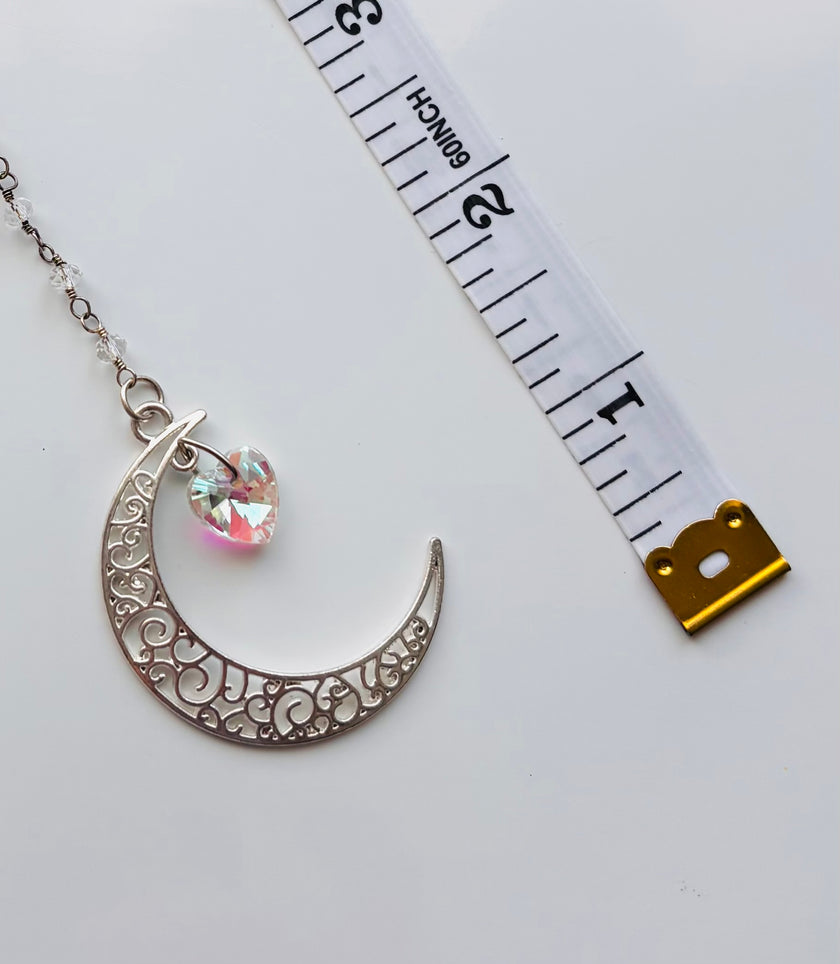 Silver crescent moon pendant with a dangling iridescent heart charm, shown next to a white measuring tape on a light gray background.