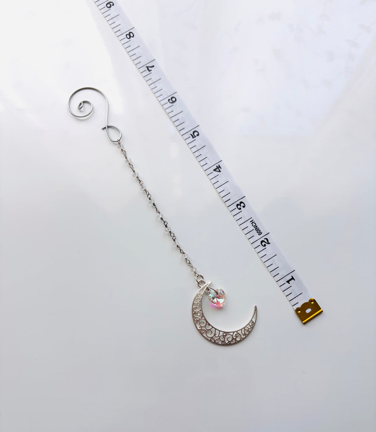 A silver spiral dangling chain and crescent moon charm embellished with beaded crystal and a small iridescent heart, shown next to a white measuring tape on a white background.