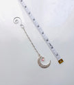 A silver spiral dangling chain and crescent moon charm embellished with beaded crystal and a small iridescent heart, shown next to a white measuring tape on a white background.