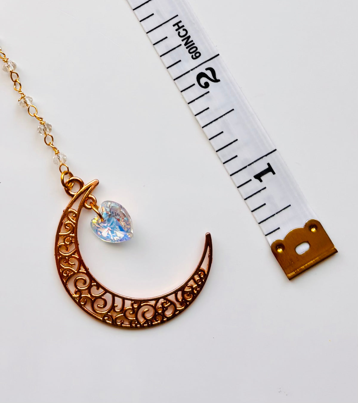 A gold crescent moon pendant with intricate scrollwork and a small iridescent heart-shaped crystal charm, shown next to a white measuring tape on a white background.