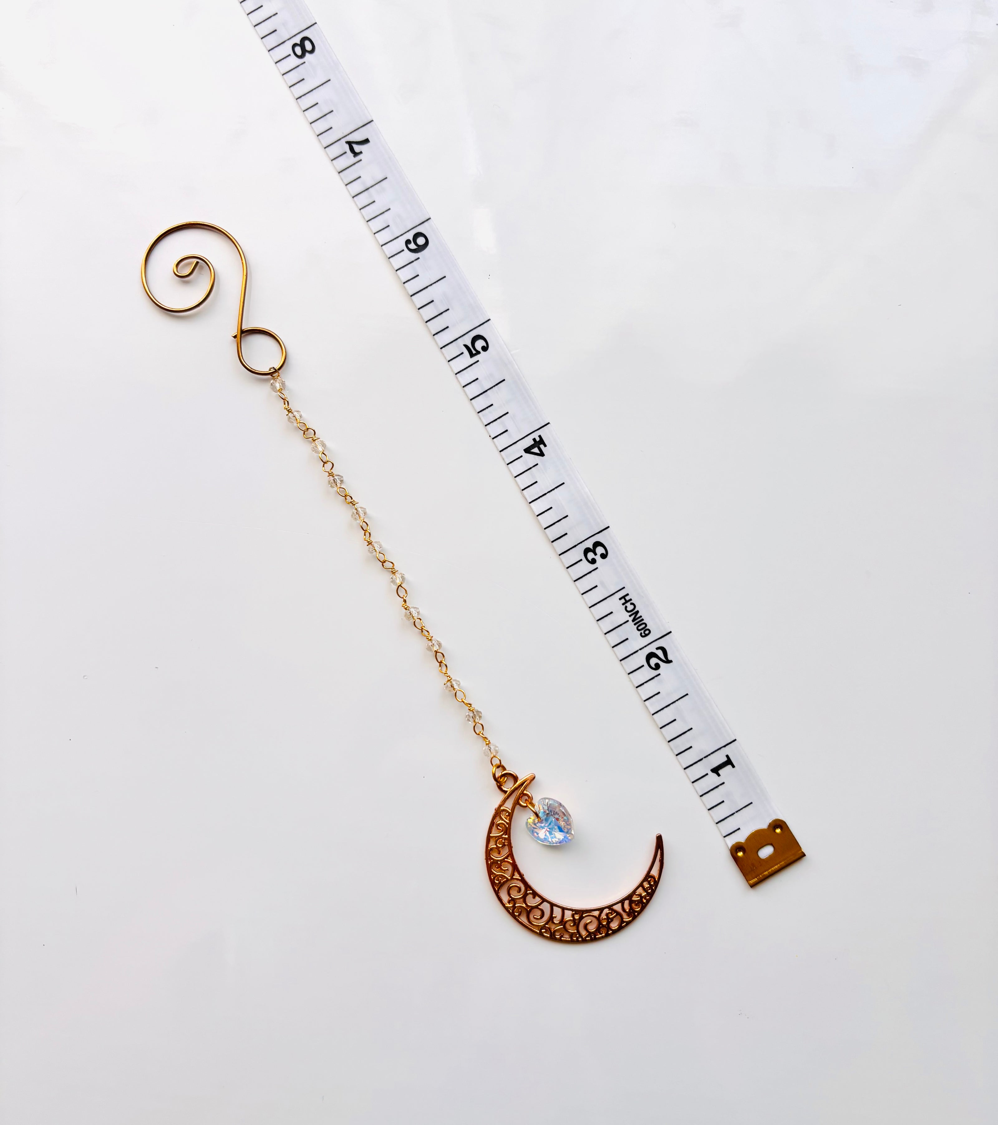 A gold crescent moon with a filigree pattern, a small aurora borealis heart faceted gemstone, and a spiral top, shown next to a white measuring tape on a white background.