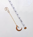 A gold crescent moon with a filigree pattern, a small aurora borealis heart faceted gemstone, and a spiral top, shown next to a white measuring tape on a white background.