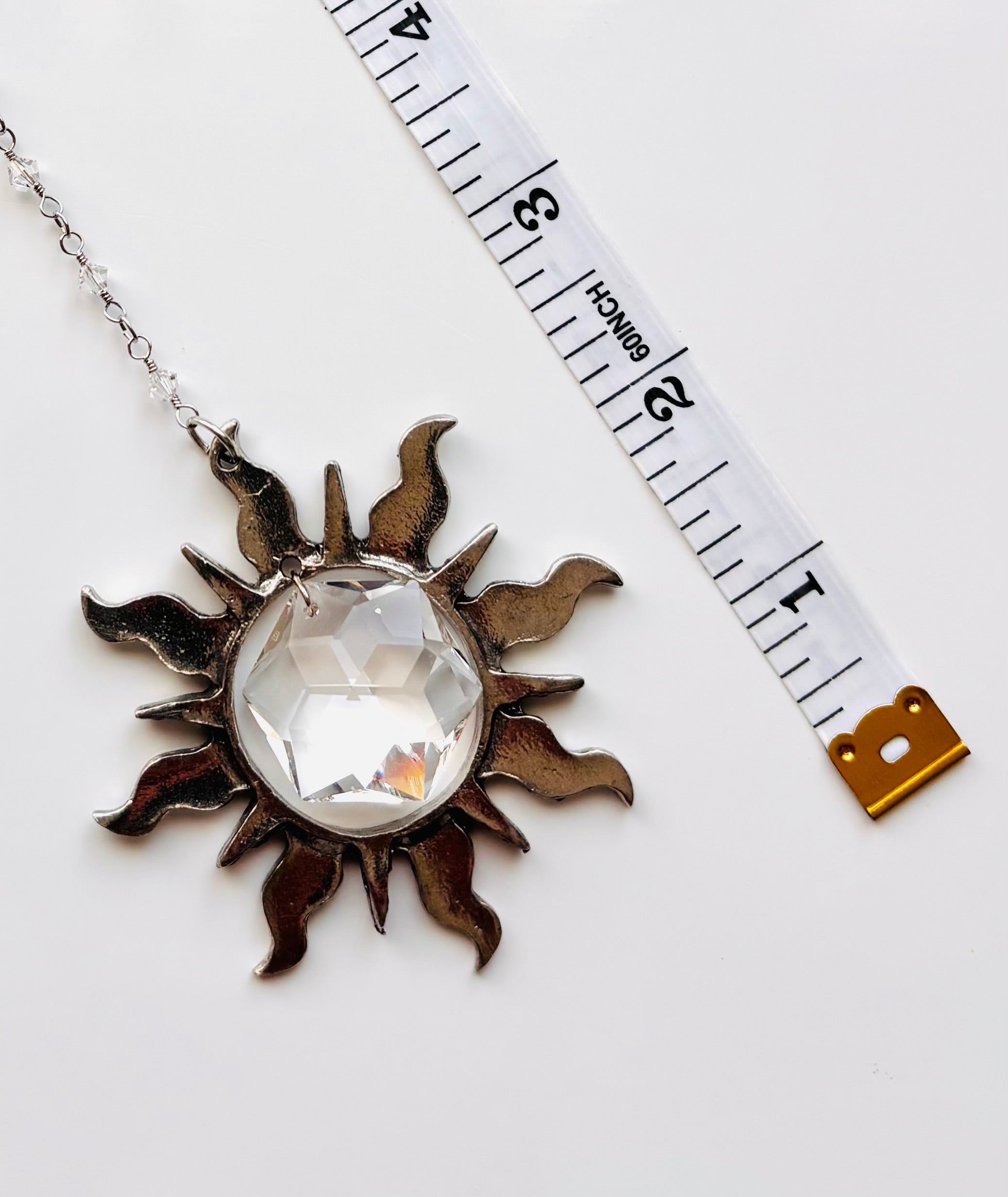 A silver sun pendant necklace with a faceted clear crystal gemstone, shown next to a white measuring tape on a white background.