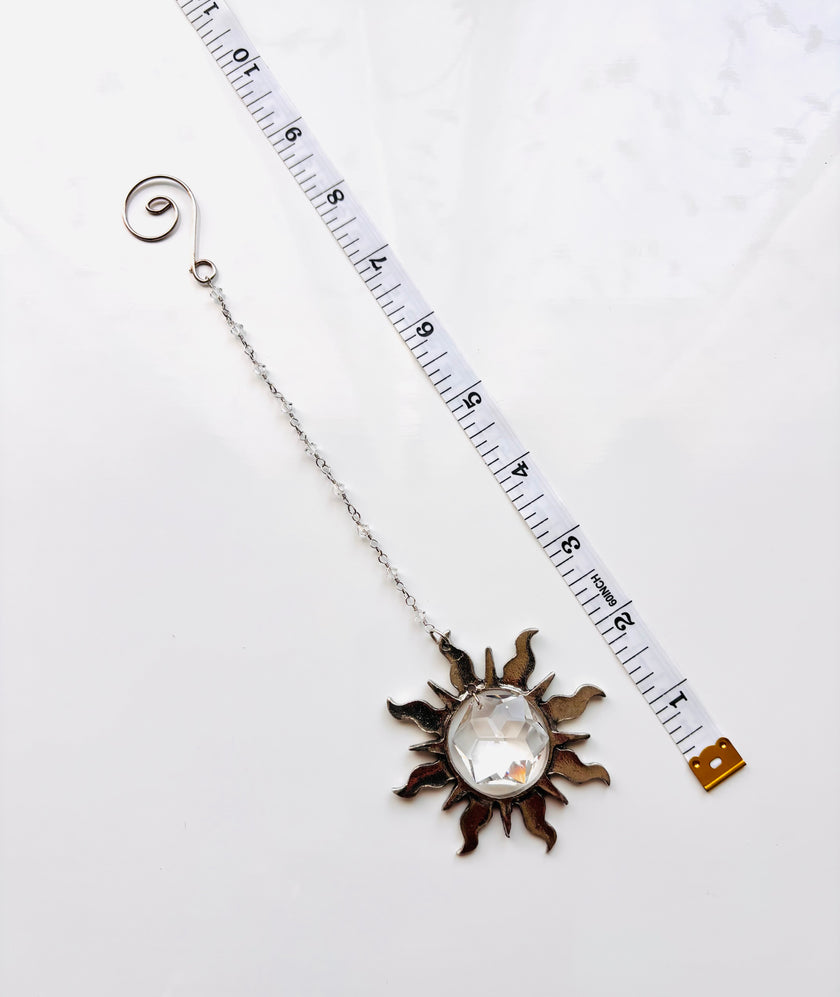 A silver-toned metal sun pendant with a clear faceted gemstone center, shown next to a white measuring tape on a white background to indicate scale.