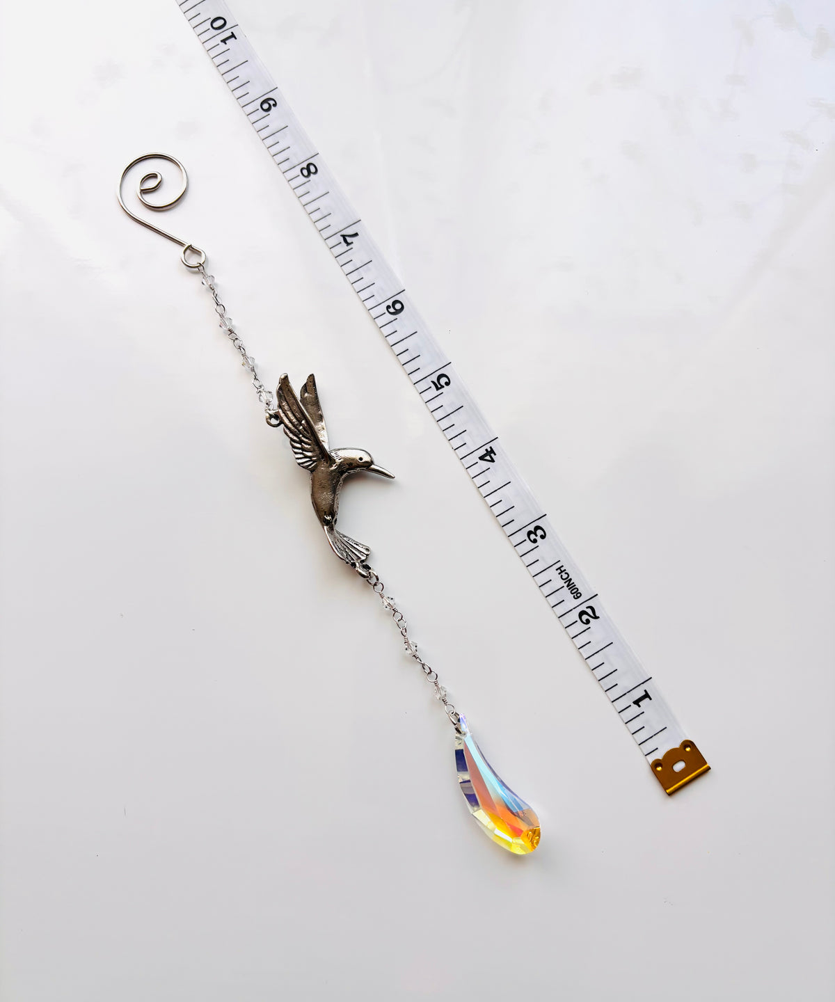 A silver-toned metal suncatcher with a hummingbird with a dangling teardrop crystal pendant, shown next to a measuring tape on a white background.