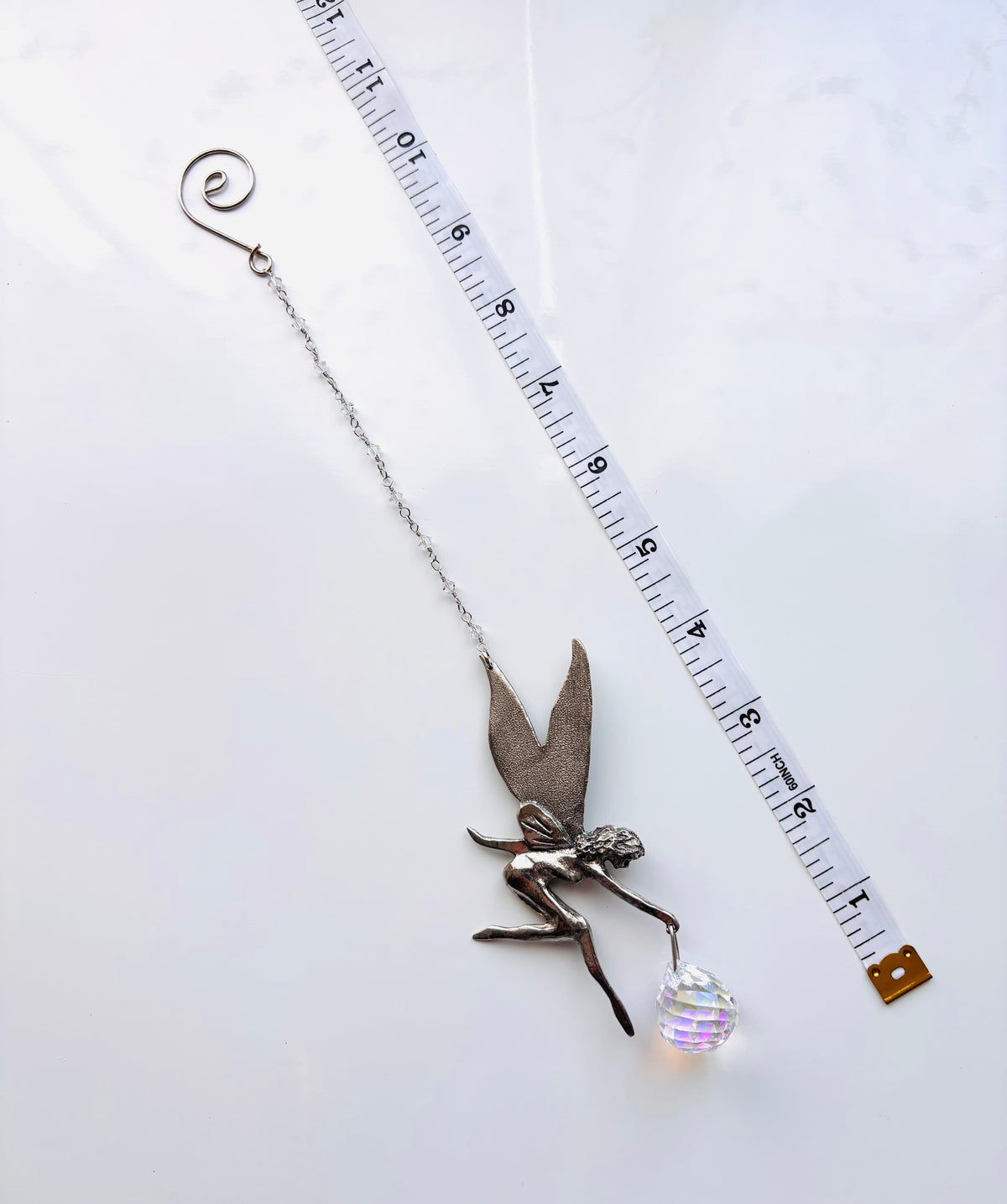 A silver fairy charm with a delicate chain, a small winged fairy pendant, and a clear faceted crystal ball, shown next to a measuring tape on a white background.