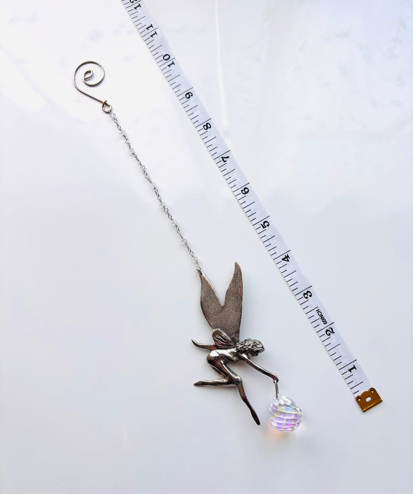 A silver fairy charm with a delicate chain, a small winged fairy pendant, and a clear faceted crystal ball, shown next to a measuring tape on a white background.