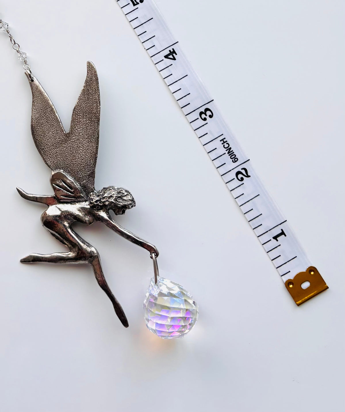 A silver fairy pendant holding a faceted iridescent crystal ball, shown next to a white measuring tape on a white background.
